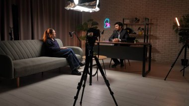 Sahne arkasında. İki konuk, mikrofon ve ışıklandırmayla donatılmış yaratıcı bir podcast stüdyosu içinde dinamik bir diyalog içerisindeler. Atmosfer heyecan ve hikaye anlatımıyla dolu.