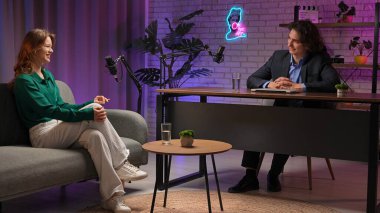 Canlı bir podcast stüdyosunda, bir sunucu rahat bir koltukta oturan bir uzmanla röportaj yapar. Atmosfer canlı, stüdyo ışıkları ve mikrofonlar ile işaretlenmiş, zengin hikaye anlatımı ve bağlantıları teşvik ediyor..