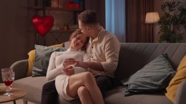 Sevgililer gününde evde hoş bir çift var. Mutlu çift, sevgililer gününü evde, kanepede kucaklaşarak kutluyor, kadın ve erkek sarılıyor ve nazikçe birbirlerine dokunuyorlar. Yavaş çekim.