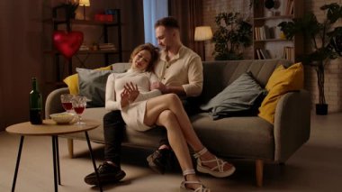 Sevgililer gününde evde hoş bir çift var. Mutlu çift, sevgililer gününü evde, kanepede kucaklaşarak kutluyor, kadın ve erkek sarılıyor ve nazikçe birbirlerine dokunuyorlar. Yavaş çekim.
