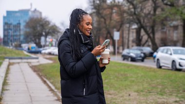Günlük rutin yaşam tarzı konsepti. Genç Afro-Amerikalı kadın sabahları şehirde günlük yürüyüşün keyfini çıkarıyor, sokakta yürüyor, akıllı telefondan mesaj atıyor, elinde kahve fincanı tutuyor..