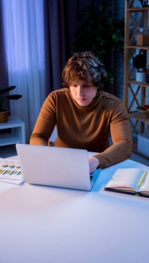 Günlük rutin yaşam tarzı konsepti. Akşamları geç saatlere kadar çalışan genç iş adamı, masada oturuyor ve kulaklık takarak dizüstü bilgisayarda çalışıyor, ekrana bakıyor, klavyede daktilo kullanıyor..