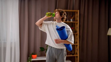 Günlük rutin yaşam tarzı konsepti. Sabahleyin yatak odasında ısınan genç adam aktif egzersizleri bitirdi. Elinde yoga minderiyle durup su içti..