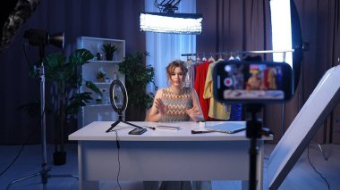 Sahne arkasında. Tutkulu bir içerik yaratıcısı, modern bir stüdyoda kayıt yaparken, moda ve güzellik hakkında uzman görüşlerini paylaşarak seyirciye bağlanır. Titreşimli arka plan onun şık giyiminin altını çiziyor..