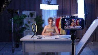 Sahne arkasında. Tutkulu bir içerik yaratıcısı, modern bir stüdyoda kayıt yaparken, moda ve güzellik hakkında uzman görüşlerini paylaşarak seyirciye bağlanır. Titreşimli arka plan onun şık giyiminin altını çiziyor..