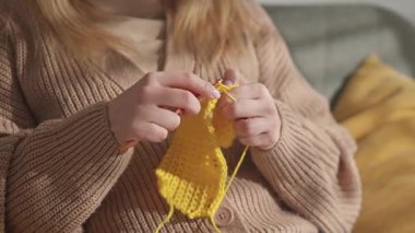 Kanca kullanarak sarı iplikle örgü ören bir kadının ellerini yakın plan çek. Arkadaki masada duran iplik ve örgü iğnelerinden oluşan bir top rahat bir atmosfer yaratıyor. Yavaş çekim.