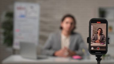 Bir içerik yaratıcısı düşüncelerini iyi aydınlatılmış bir stüdyoda paylaşır, bir masada dizüstü bilgisayarla oturur ve bir akıllı telefon aracılığıyla izleyicilerle iletişim kurar..
