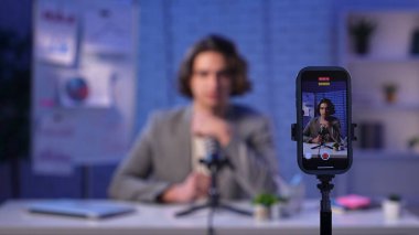 Parlak ve şık bir podcast stüdyosunda, içerik yaratıcısı güven içinde masaya oturur, röportaj için hazırdır. Bir kamera değiş tokuşu kaydeder, yaratıcılık sürecini vurgular..