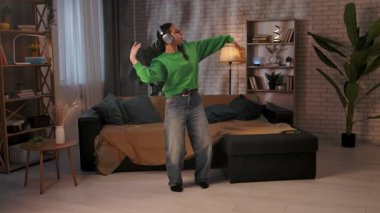 Yeşil kazaklı ve kotlu genç bir kadın sıcak ışıklandırması ve şık dekoruyla modern bir oturma odasında modern dans figürleri sergiliyor. Yavaş çekim.