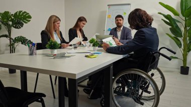 Aralarında tekerlekli sandalyedeki bir profesyonelin de bulunduğu çeşitli bir iş ekibi modern bir ofiste strateji toplantısı yaptı. İşbirliği, eşitlik ve işyeri dahil etme kavramı.