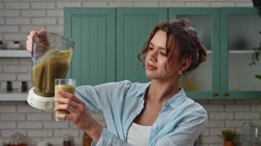 Ev yemeği ve sağlıklı beslenme konsepti. Mutfaktaki masada oturan genç bir kadın sağlıklı kahvaltı için smoothie hazırlıyor, bardağa taze meyve suyu dolduruyor ve bir yudum alıyor, tadını çıkarıyor. Yavaş çekim