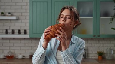 Ev yemeği ve sağlıklı beslenme konsepti. Mutfaktaki masada oturan ve sağlıklı bir kahvaltı hazırlayan genç kadın, taze pişmiş kruvasan yiyip tadını çıkarırken yakın plan çekilmiş. Yavaş çekim