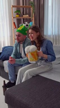 Hafta sonları birlikte birkaç boş zaman geçiririz. Çift evdeki kanepede dinleniyor, erkek ve kadın futbol hayranları TV 'de maç izliyor, takımı alkışlamayı bekliyor, mutlu bir şarap cümlesi. Dikey video.