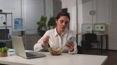 İş hayatı konsepti. İş kadını ofiste masa başında yemek dağıtımı yapıyor sağlıklı salata yiyen ve akıllı telefondan sosyal medyayı karıştıran bir kadın. Yavaş çekim