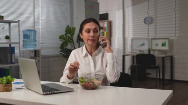 İş hayatı konsepti. İş kadını ofiste masa başında yemek dağıtıyor, sağlıklı salata yiyor ve iş arkadaşıyla telefonda dedikodu yapıyor. Yavaş çekim