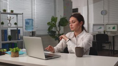 İş hayatı konsepti. İş kadını ofiste masa başında yemek servisi yapıyor, bilgisayarını kapatmayı bitiriyor, yorgun bir ifade takınıyor, sandalyeye oturup kahve içiyor. Yavaş çekim