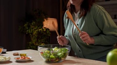 Sağlıklı beslenme konsepti. Mutfaktaki masada oturan kadın akşam yemeği için sağlıklı sebze salatası hazırlıyor kulaklıkla müzik dinliyor spatula ile dans ediyor ve içindekileri kaseye karıştırıyor. Yavaş çekim