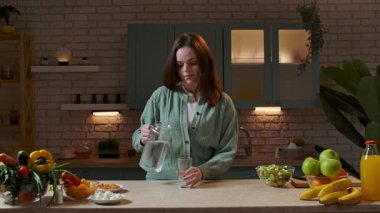 Sağlıklı beslenme konsepti. Mutfaktaki masada akşam yemeği için sağlıklı sebze salatası hazırlayan kadın mutfağa gelir, bardağa su koyar ve içer. Yavaş çekim