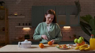 Sağlıklı beslenme konsepti. Mutfaktaki masada oturan kadın akşam yemeği için sağlıklı sebze salatası hazırlıyor, güvertede taze portakallar kesiyor meyve suyu için malzemeler hazırlıyor. Yavaş çekim