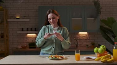 Sağlıklı beslenme konsepti. Mutfaktaki masada oturan kadın akşam yemeği için sağlıklı sebze salatası hazırlıyor, tabağın resmini salatayla çekiyor ve akıllı telefondan selfie çekiyor. Yavaş çekim