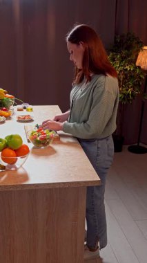 Sağlıklı beslenme konsepti. Mutfaktaki masada oturan kadın akşam yemeği için sağlıklı sebze salatası hazırlıyor, güvertede taze salatalık kesiyor ve kaseye ekliyor. Dikey video