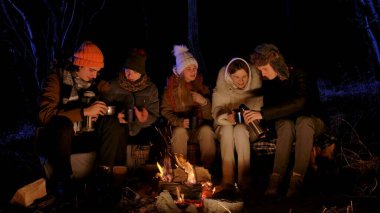 Bir grup genç arkadaş sıcak kış kıyafetleri içinde gece parlayan bir kamp ateşinin etrafında oturuyorlar. Metal bardak ve termos tutuyorlar, sıcak içecekleri paylaşıyorlar ve birlikte dışarıda eğleniyorlar..