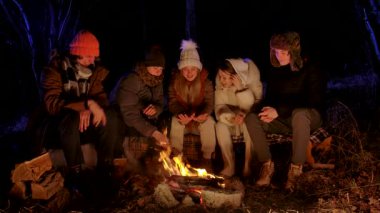 Sıcak kış kıyafetleri içinde bir grup arkadaş karanlık bir ormanda şenlik ateşinin etrafında oturmuş, hikayelerini paylaşıyor ve soğuk bir gecede ateşin sıcaklığının tadını çıkarıyorlar..