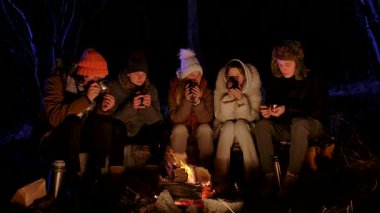 Bir grup genç arkadaş sıcak kış kıyafetleri içinde gece parlayan bir kamp ateşinin etrafında oturuyorlar. Metal bardak ve termos tutuyorlar, sıcak içecekleri paylaşıyorlar ve birlikte dışarıda eğleniyorlar..