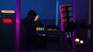 Neon ışıklı bir kayıt stüdyosunda profesyonel bir podcast mikrofon. Bulanık arkaplanda ses mühendisi ve parlayan ses ölçerler var. Modern bir yayın atmosferi yaratıyor..