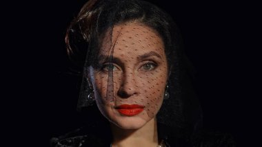 Stüdyoda profesyonel model. Siyah arka planda izole edilmiş kameraya poz veren eski moda elbiseli kadın oyuncu, peçe takan ve doğrudan bakan kadın..