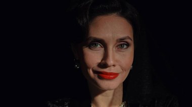 Stüdyoda profesyonel model. Siyah arka planda izole edilmiş fotoğraf makinesine poz veren eski moda elbiseli kadın oyuncu, gülümseyen ve doğrudan bakan kadının yakın çekimi..