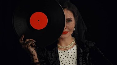 Stüdyoda profesyonel model. Siyah arka planda izole edilmiş kameraya poz veren eski moda saç stili ve makyajlı kadın oyuncu, kameraya poz veriyor, yüzünün yarısını eski plaklarla gizliyor..