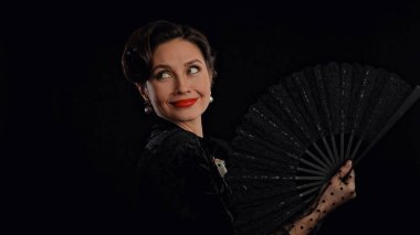 Stüdyoda profesyonel model. Siyah arka planda izole edilmiş kameraya poz veren eski moda elbiseli kadın oyuncu, dantel pervaneyle gülümseyen ve poz veren kadın..