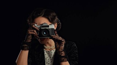 Stüdyoda profesyonel model. Siyah arka planda izole edilmiş kameraya poz veren eski moda saç stili ve makyajlı kadın oyuncu, gülümseyen ve eski film kamerasıyla poz veren kadın..