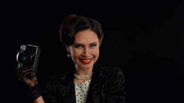 Stüdyoda profesyonel model. Siyah arka planda izole edilmiş kameraya poz veren eski moda saç stili ve makyajlı kadın oyuncu, gülümseyen ve eski film kamerasıyla poz veren kadın..