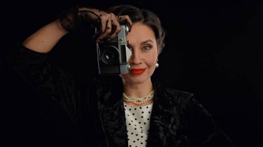 Stüdyoda profesyonel model. Siyah arka planda izole edilmiş kameraya poz veren eski moda saç stili ve makyajlı kadın oyuncu, gülümseyen ve eski film kamerasıyla poz veren kadın..