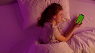 Yatak odasındaki kız. Yatakta, üzerinde pembe neon ışıklarla kaplı bir battaniyeyle uzanmış genç bir kadın elinde krom renkli yeşil ekran arka planlı bir akıllı telefon tutuyordu. Üst görünüm.