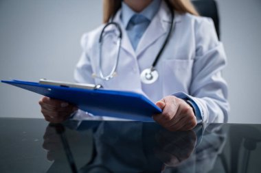 Ofiste çalışan bir tıp uzmanı. Beyaz önlüklü kadın klinikteki çalışma masasında oturmuş elinde tıbbi evraklarla mavi bir dosya tutuyor..