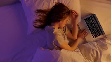 Yatak odasındaki kız. Mor neon ışıkla kaplı yastığın üzerinde uyuyan genç bir kadın, dizüstü bilgisayarda el ele tutuşarak video izlerken uyuyakaldı. Üst görünüm.