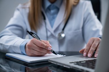 Ofiste çalışan bir tıp uzmanı. Beyaz önlüklü kadın doktor klinik odasında masa başında oturuyor, dizüstü bilgisayar kullanıyor, tıbbi evraklarla uğraşıyor, deftere yazı yazıyor..