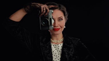 Stüdyoda profesyonel model. Saç stili ve makyajlı kadın oyuncu siyah arka planda izole edilmiş, eski fotoğraf makinesinde fotoğraf çekiyor ve gülümsüyor. Yavaş çekim.