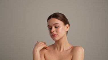 Doğal güzellik tedavisi ve cilt bakımı. Kameraya poz veren genç bir kadın neşeli bir şekilde gülümsüyor pürüzsüz tenine dokunuyor, beyaz arka planda izole edilmiş. Yavaş çekim.