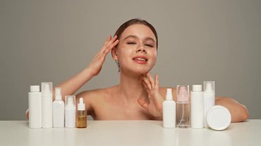 Doğal güzellik tedavisi ve cilt bakımı. Kameraya gülümseyen genç bir kadın kozmetik ürünlerle masada oturuyor, yüzüne su püskürtmeyi seçiyor, beyaz arka planda izole edilmiş..