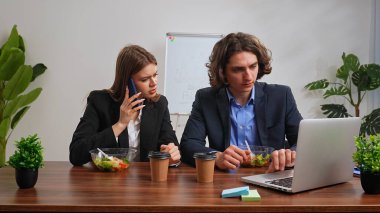 İş hayatı konsepti. Ofiste masa başında oturan, yemek dağıtan, salata yiyen, akıllı telefondan konuşan, dizüstü bilgisayar kullanan kadın ve erkeklerden oluşan bir iş ekibi..