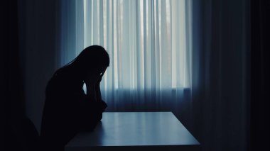 Gölgeli bir figür pencerenin kenarında tek başına oturur loş bir odada yalnızlık ve içgözlem hissi taşır..
