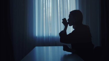 Astım spreyi soluyan bir kadının koyu renk silüeti. Akşamları karanlıkta oturan ve öksürük, alerji ya da astım için ilaç alan bir kadın..