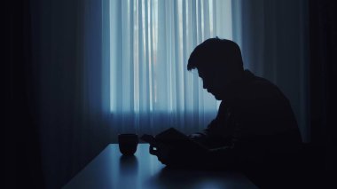 Pencerenin yanında kitap okuyan bir adamın silüeti masanın üzerinde bir fincan ile, huzur dolu bir yalnızlığı çağrıştırıyor..