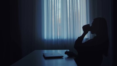 Masada dizüstü bilgisayarı olan ve bir fincan kahvenin tadını çıkaran yalnız bir insan silueti..