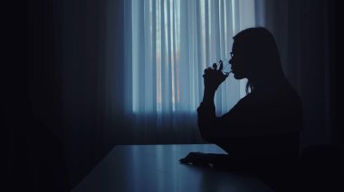 Bir kadın bardaktan yudumluyor, silueti yumuşak pencere ışığıyla çerçevelenmiş, sakin ve düşünceli bir atmosfer yaratıyor..