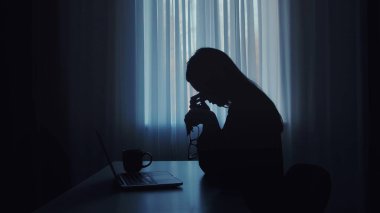 Masada oturan bir kadının silüeti, dizüstü bilgisayarın önünde, yanında kahve fincanı olan....